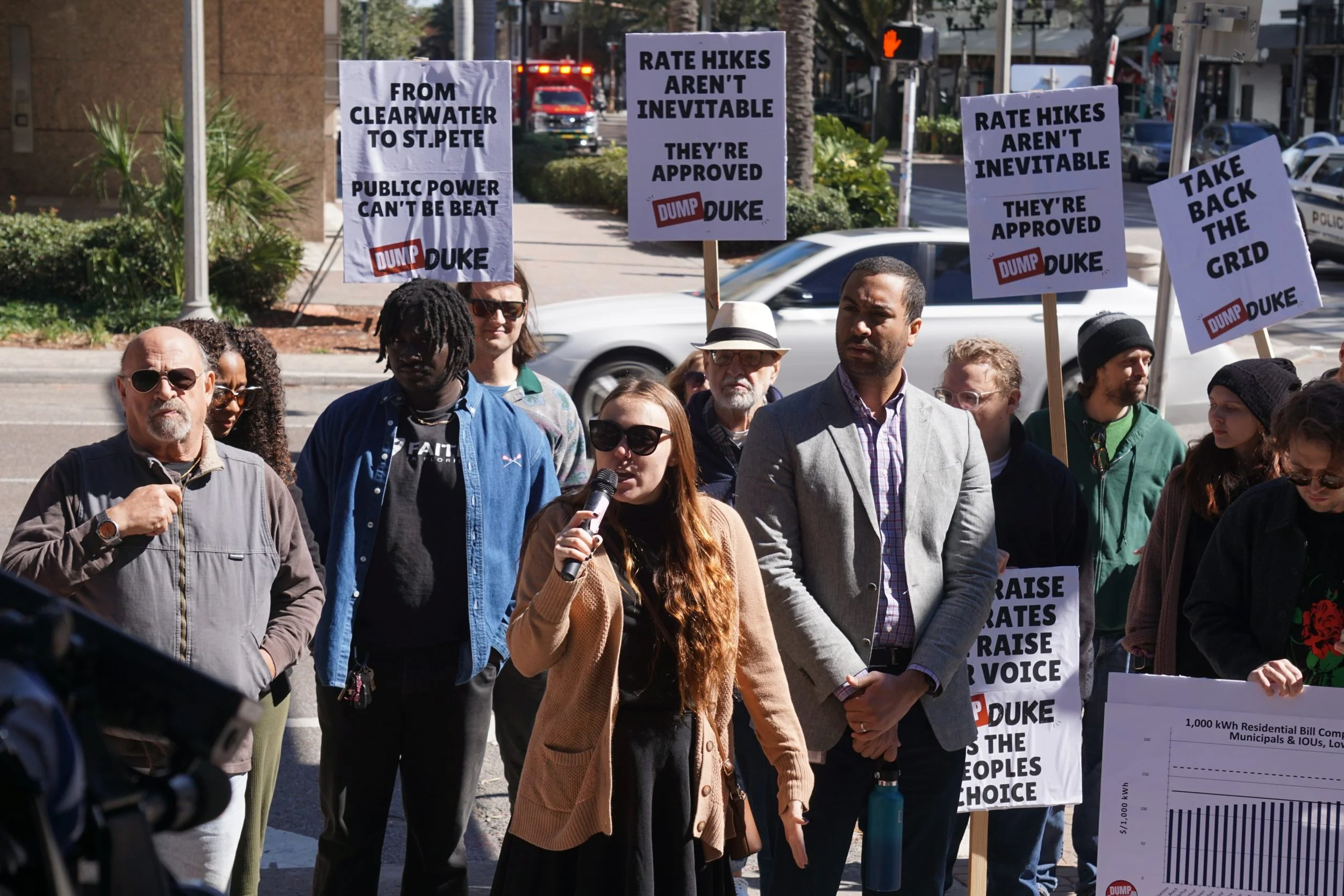 A group of people participating in a protest or rally on a city street, holding signs with messages supporting public power and criticizing rate hikes, with cars and buildings visible in the background.