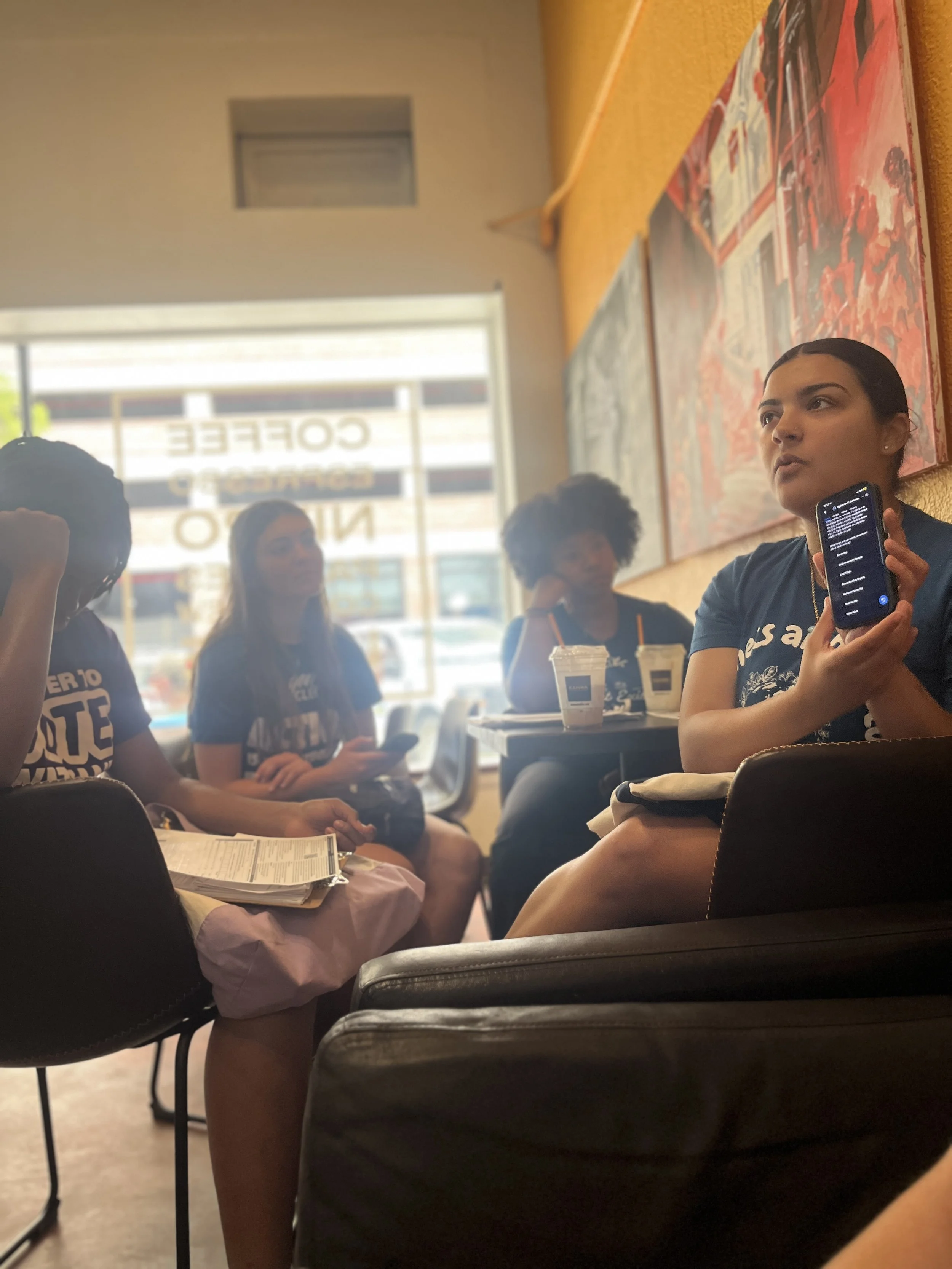 Group of people sitting at a table inside a cafe, some holding phones, with drinks on the table, view through window with sign reading 'Coffee' and other letters.