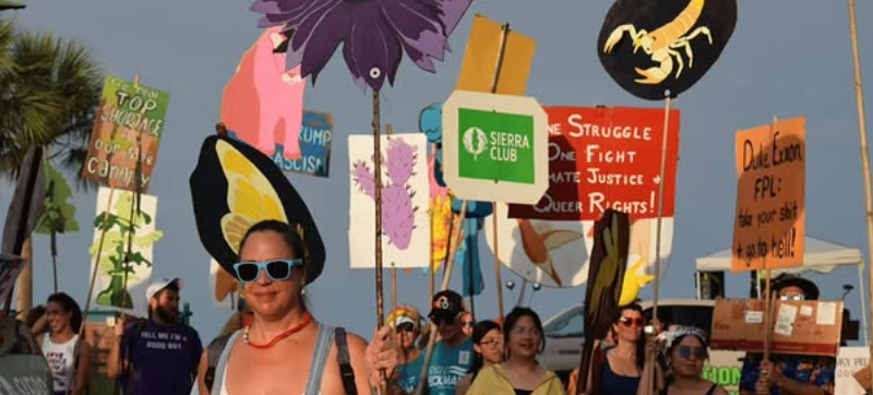 Protest crowd holding signs and banners advocating for climate action, social justice, and LGBTQ+ rights during daylight.