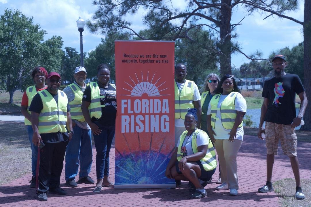 Group of people wearing yellow safety vests standing outdoors around a Florida Rising banner on a brick-paved area, with trees and a lamppost in the background.