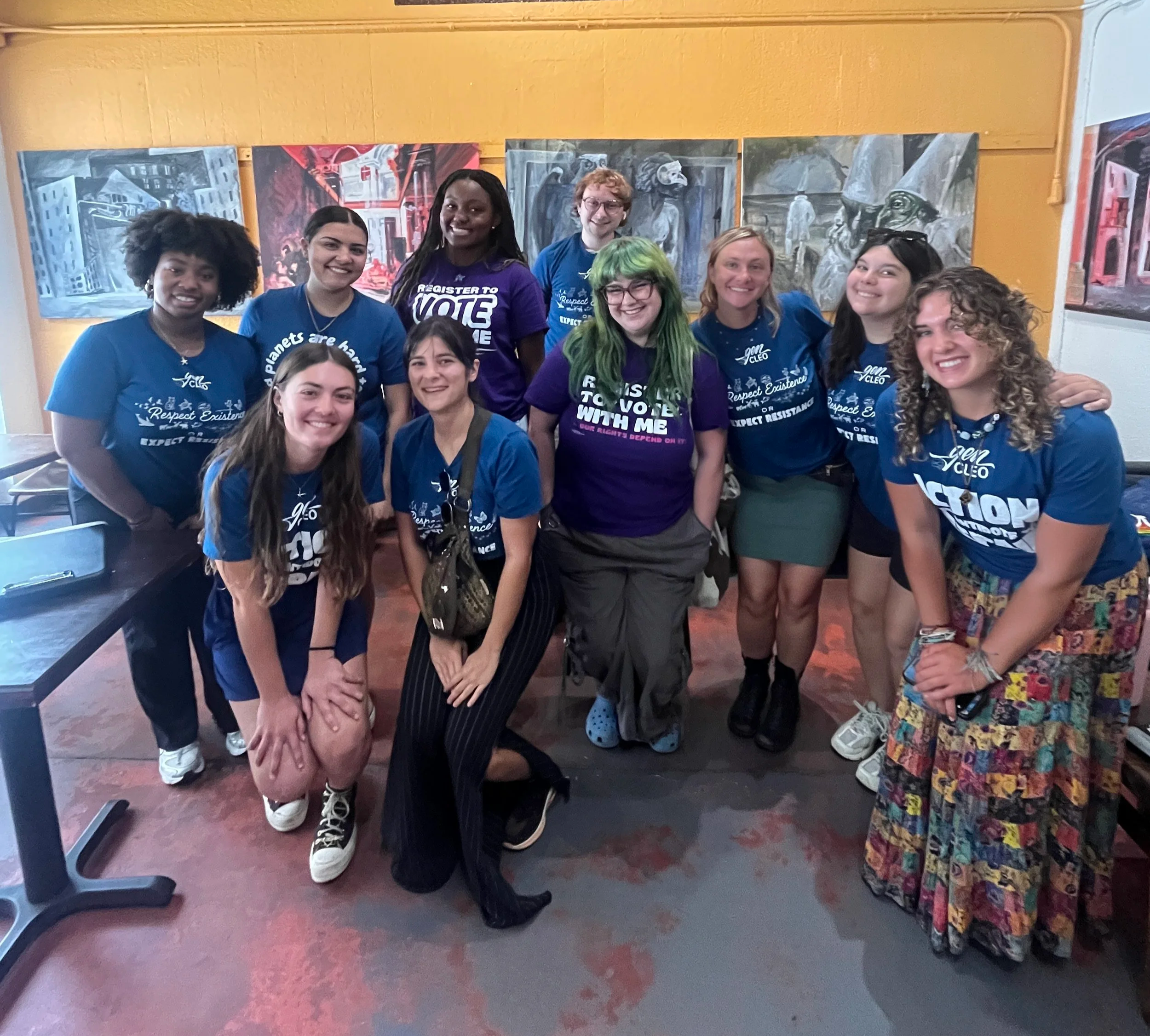 A group posing together indoors in front of painted murals, smiling at the camera. They are wearing matching blue or purple t-shirts with slogans promoting respect and voting, and some are wearing glasses. The background features artistic murals of buildings and abstract scenes.