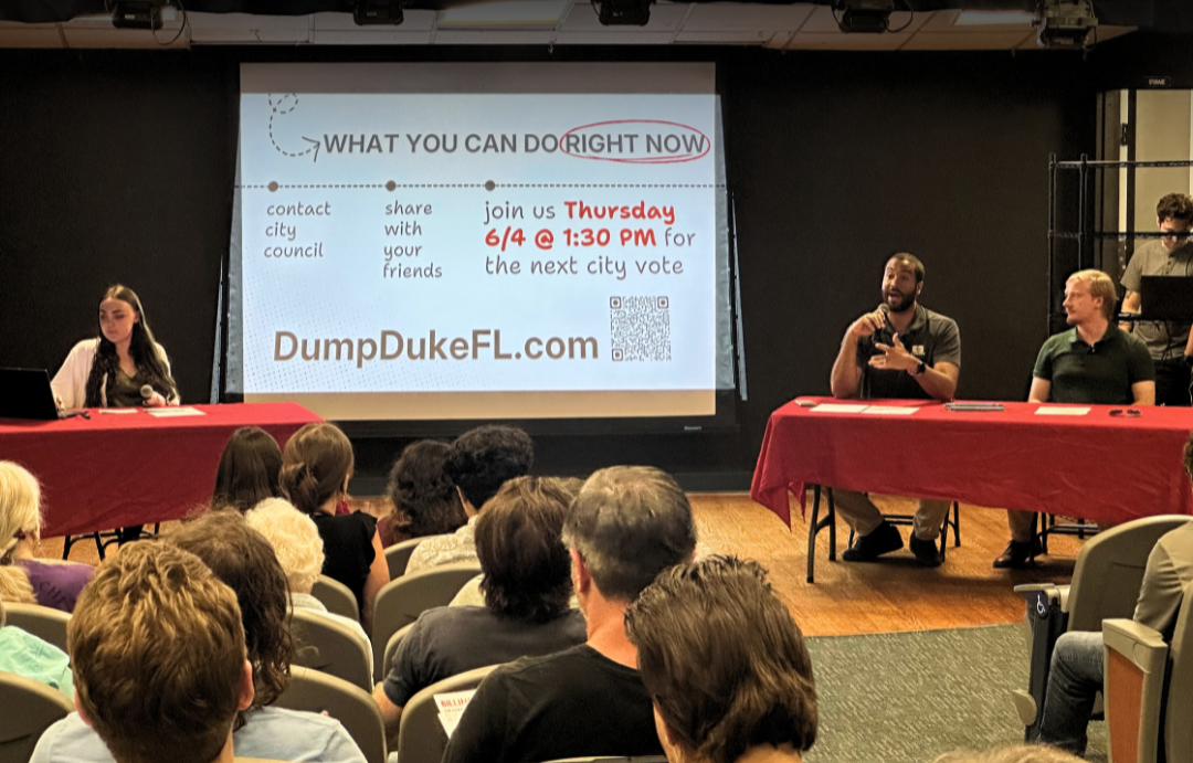 People attending a panel discussion about city voting, with a presentation on a large screen that mentions contacting city council and joining a city vote on Thursday, June 4th at 1:30 PM, with a website link DumpDukeFL.com.