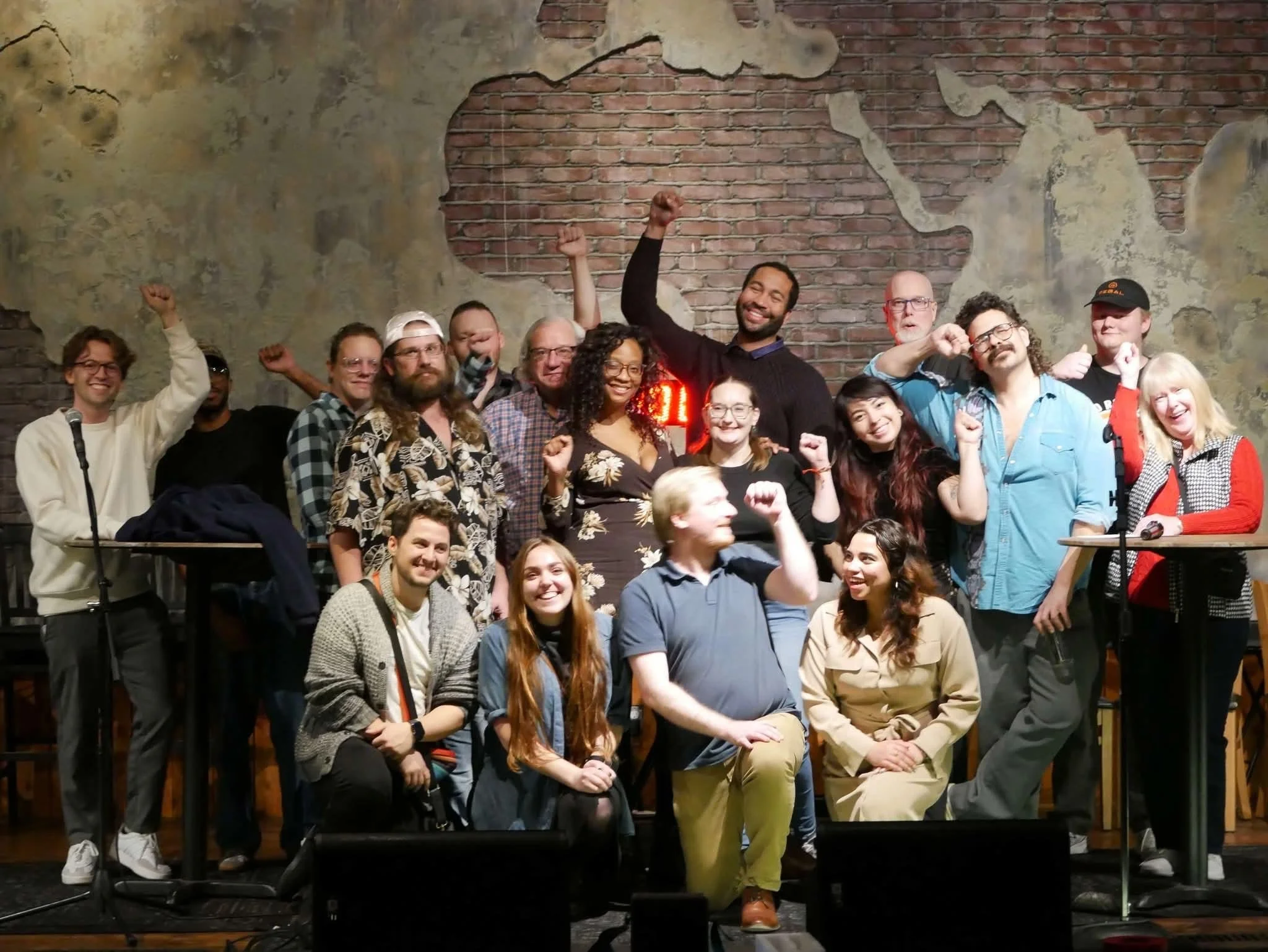 A diverse group of people standing and posing together inside a room with exposed brick and plaster walls, some smiling and raising their fists in celebration.