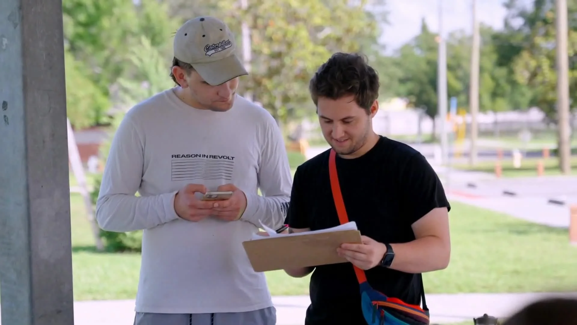 Two young men outdoors at a park or walkway, one holding a phone and the other with a clipboard, both looking at the paper, engaged in conversation.