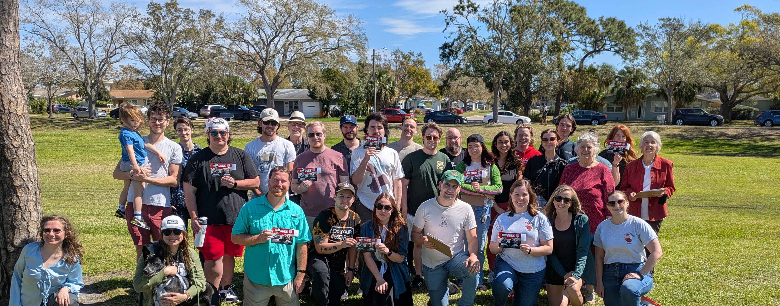 A diverse group of people posing outdoors on a sunny day in a park, some holding signs that say 'DUKE.' They are standing on green grass with trees and houses in the background.