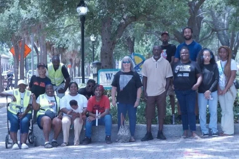 Group of diverse people, including children and adults, posing for a photo in a park with trees and a sign in the background.