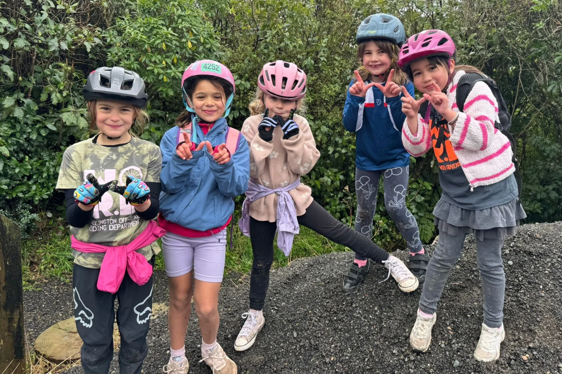 Five young girls wearing helmets and outdoor clothing, standing on a trail with greenery in the background. They are smiling and making peace signs or pointing gestures, enjoying an outdoor adventure.