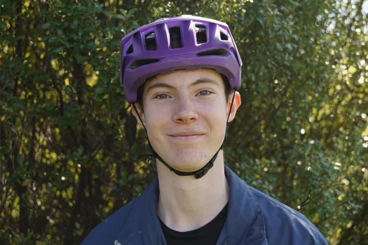 Young man wearing a purple bicycle helmet outdoors with green trees in the background