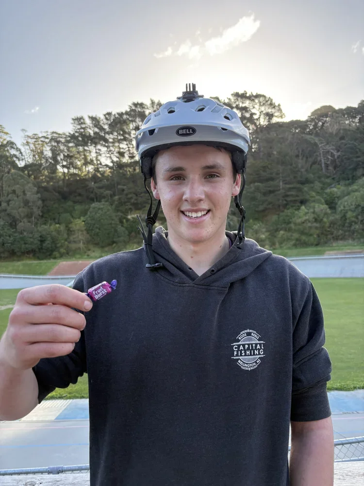 A young man wearing a white bicycle helmet and a black hoodie with "Capital Fishing" logo, smiling and holding a small purple candy in an outdoor sports field with trees and sky in the background.