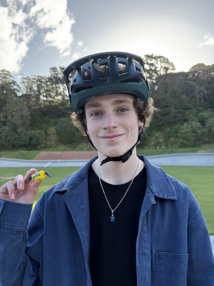 Young man with curly hair wearing a helmet, black shirt, and blue jacket, holding a yellow snack in an outdoor park with trees, grass, and a track in the background.