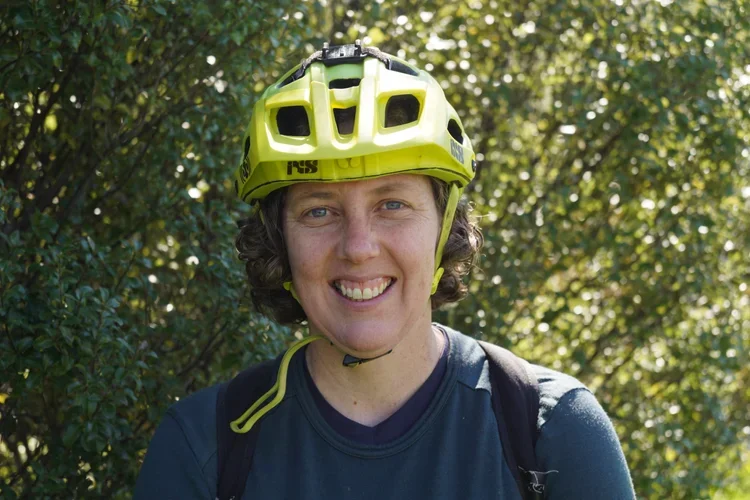 Woman wearing a yellow biking helmet and navy shirt, smiling outdoors with greenery in the background.