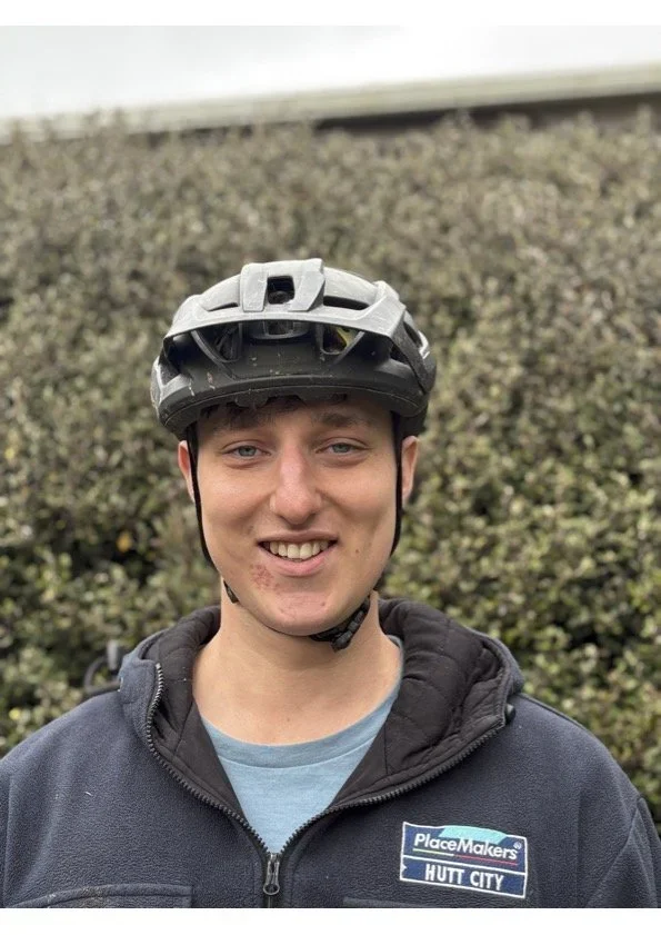 A young man wearing a gray bicycle helmet and a black jacket smiling outdoors in front of a shrubbery.