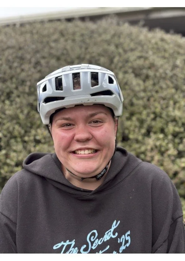Smiling person wearing a black hoodie and a gray bike helmet outside with bushes in the background.