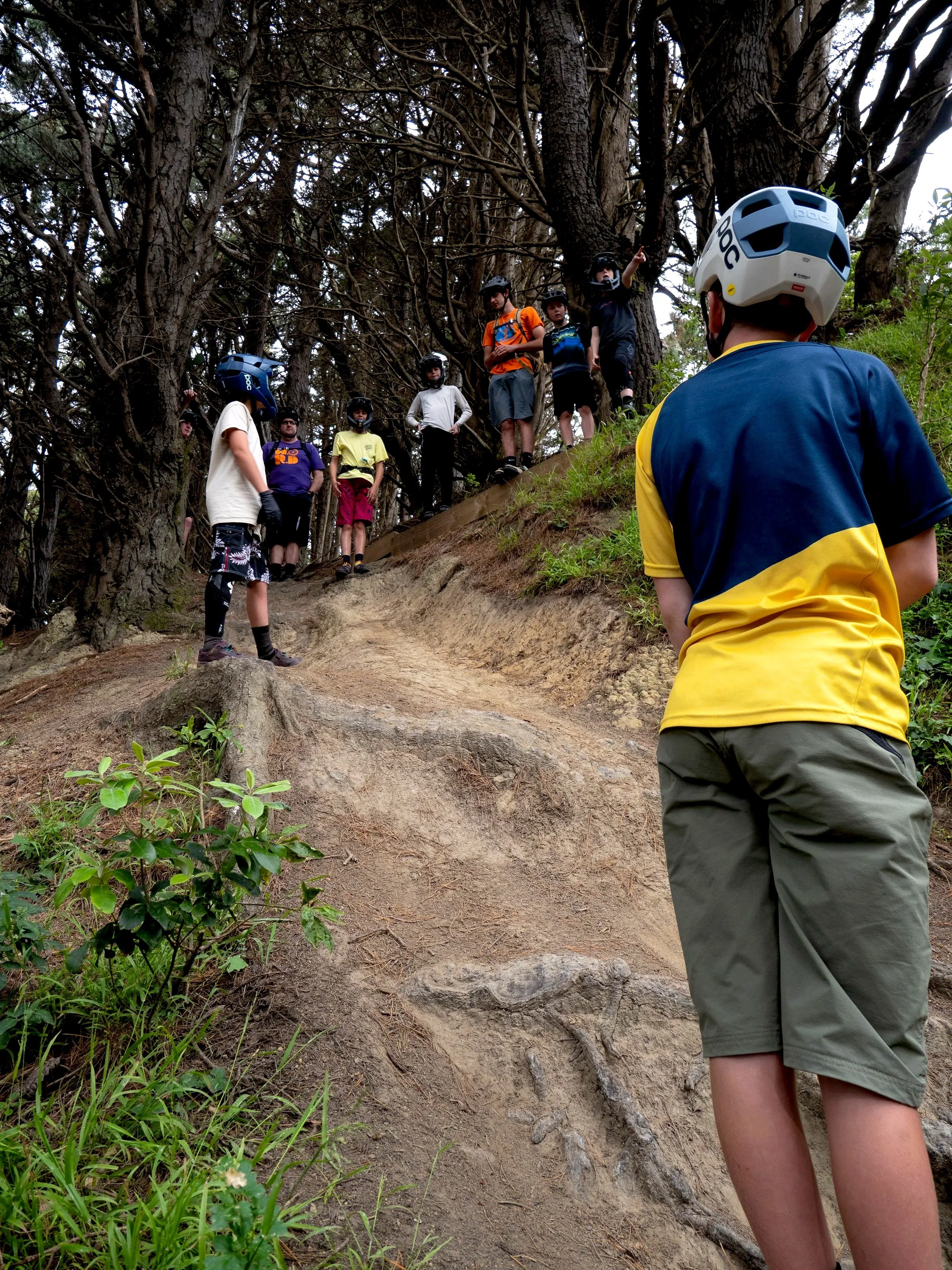 A group of children and adults with helmets standing on a dirt trail in a dense forest, with some children walking on a narrow ridge and one person pointing ahead.