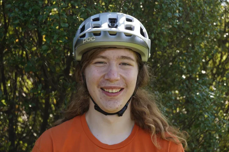 A smiling person with long red hair wearing an orange shirt and a helmet outdoors.