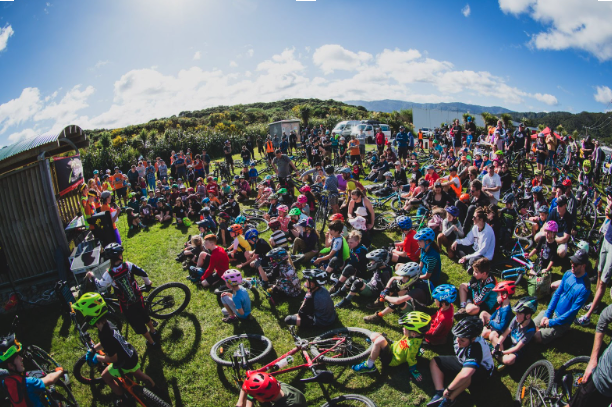 Large outdoor gathering of children and adults, many wearing helmets, with bicycles on a grassy field under a blue sky.