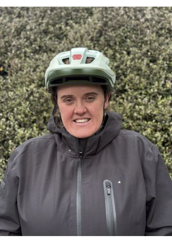 Woman wearing a bicycle helmet and a black jacket, standing outdoors in front of greenery.