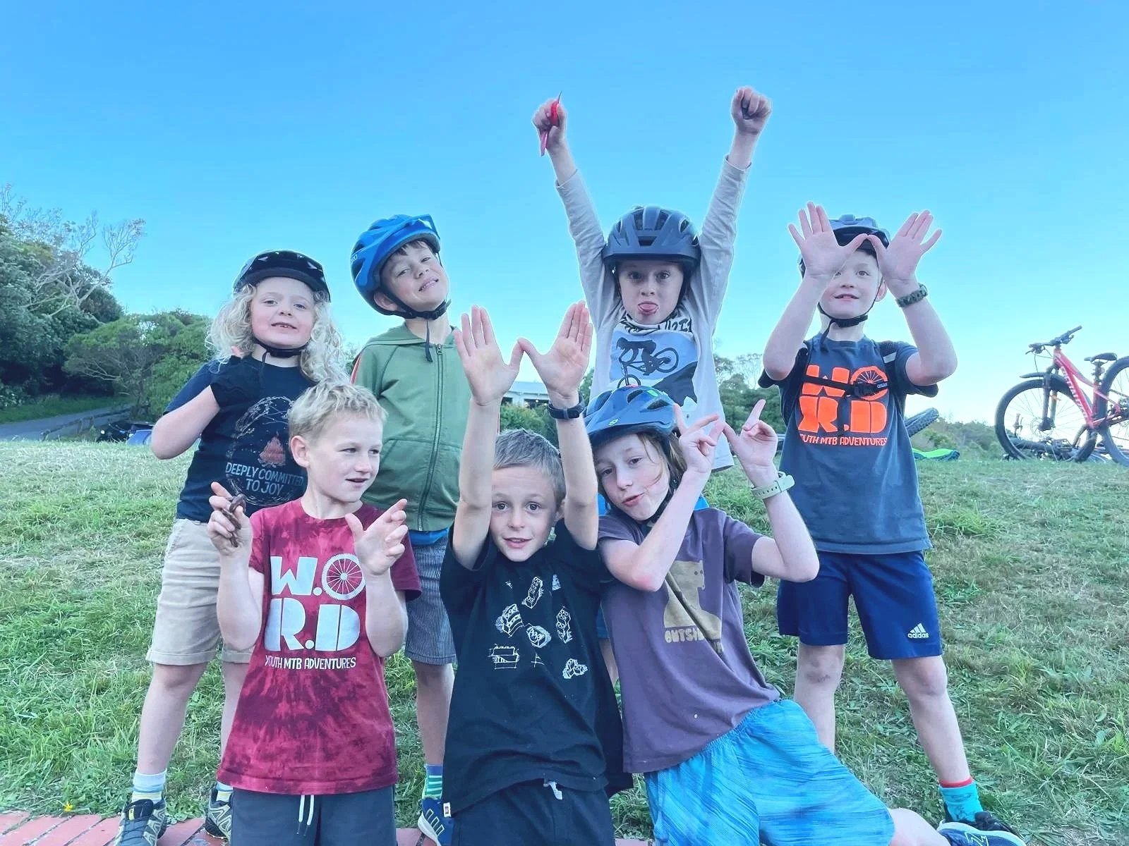Group of seven children outdoors on a grassy area, some wearing bike helmets, making various gestures and poses, with bicycles in the background.