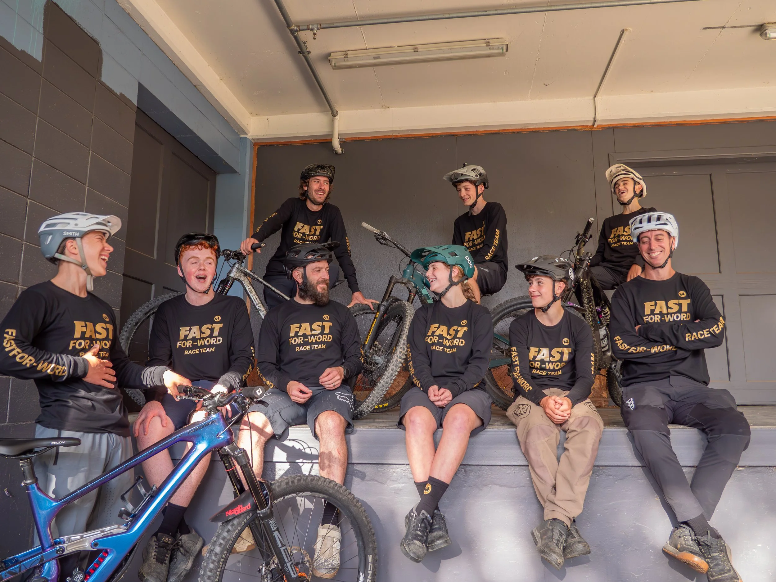 A group of eight people with bikes, wearing black shirts that say 'Fast for Word Race Team', are sitting and standing in front of a dark indoor wall, smiling and laughing.