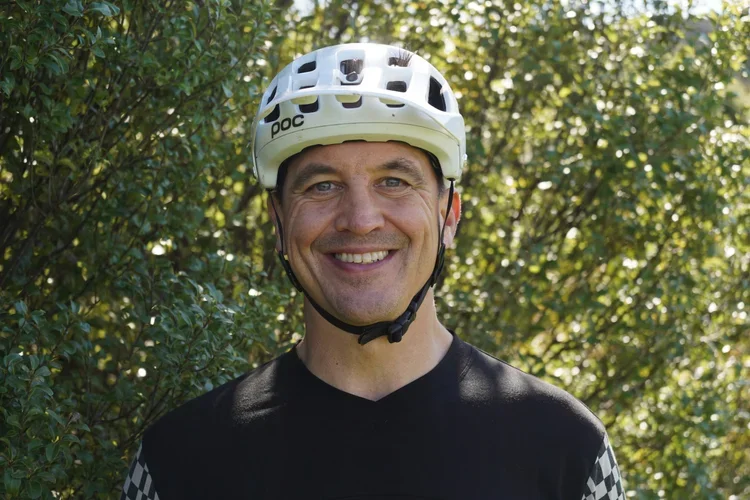 A smiling man in a black cycling shirt and white helmet outdoors in front of green foliage.