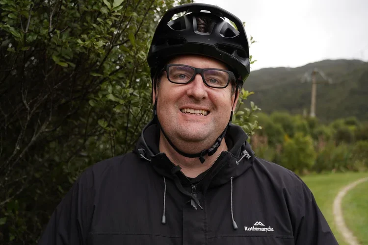 A smiling man wearing a black bicycle helmet and glasses, standing outdoors with trees and a hilly landscape in the background.