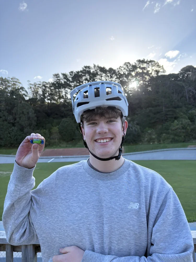 A young man wearing a white bicycle helmet and a grey sweatshirt standing outdoors on a sunny day, holding a small object in his right hand, smiling at the camera with a grassy field, trees, and a blue sky in the background.