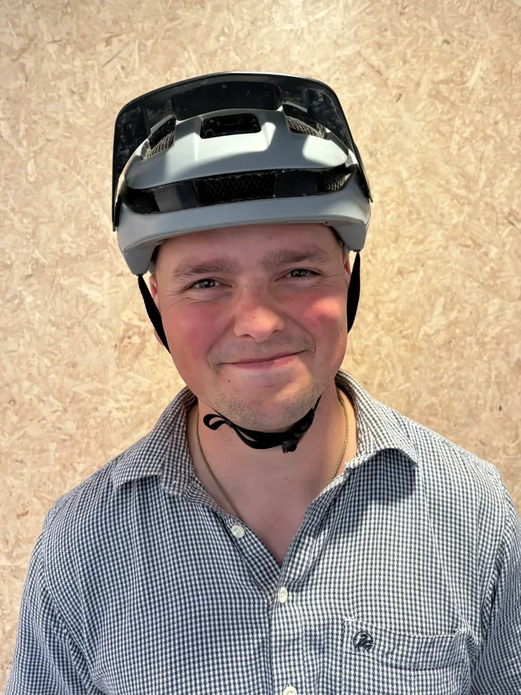 A young man smiling, wearing a bicycle helmet and a checkered button-up shirt, standing against a beige textured wall.