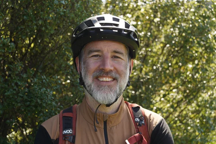 A smiling man wearing a black bicycle helmet, brown jacket, and a red backpack outdoors with green foliage in the background.