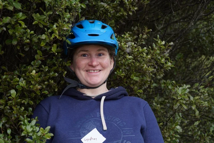 A person with short hair wearing a blue helmet and a navy blue hoodie, smiling and standing among green bushes.