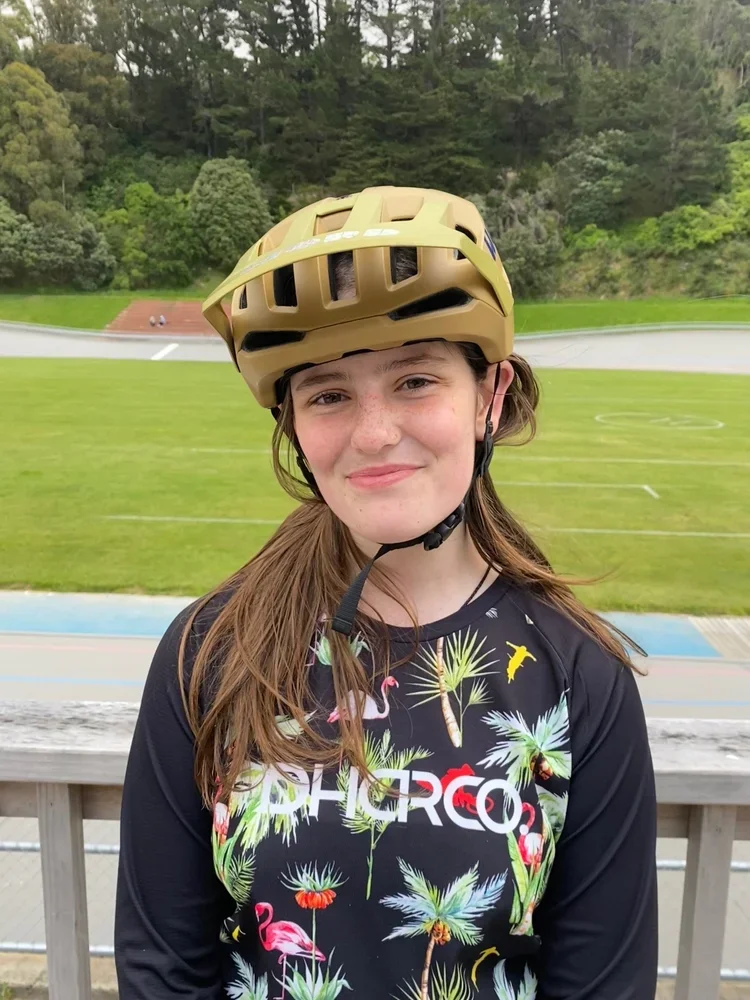 Girl wearing a tan helmet and a black sports shirt with colorful flamingos and palm trees, standing outdoors on a wooden railing with a velodrome track and greenery in the background.