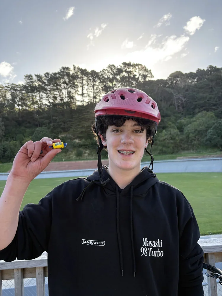 A teenage boy wearing a red bicycle helmet and black hoodie with 'Masashi 98 Turbo' written on it, holds up a small yellow and blue object while smiling outdoors with a field, trees, and a cloudy sky in the background.