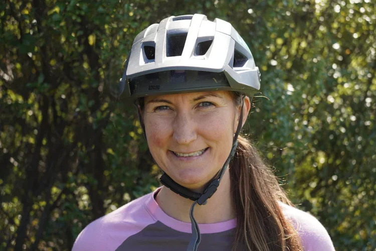 A young woman outdoors wearing a bike helmet and a pink and gray cycling jersey, smiling at the camera.