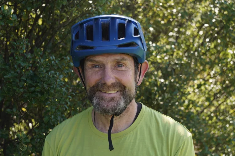 A man with a beard wearing a blue bicycle helmet and a yellow shirt, standing outdoors in front of green trees.