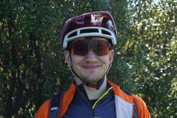 A man wearing a maroon bicycle helmet, sunglasses, and an orange and black cycling jacket, standing outdoors with green foliage in the background.