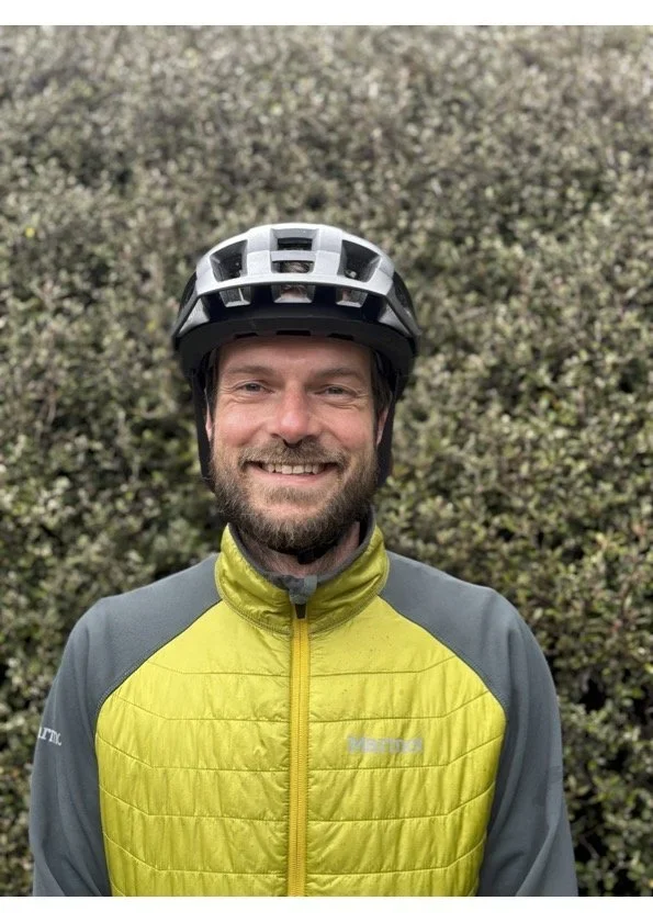 A man smiling wearing a black helmet and a yellow and gray outdoor jacket, standing in front of a bushy background.