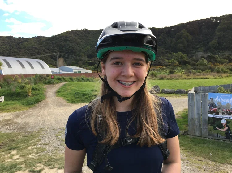 A young girl wearing a bike helmet and riding gear outside, smiling at the camera with a green hilly landscape and farm buildings in the background.