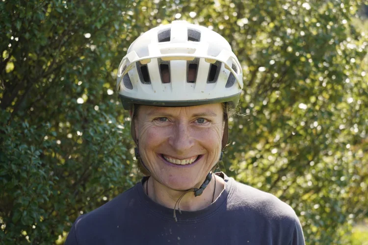 A smiling man wearing a white bicycle helmet outdoors with green trees and foliage in the background.