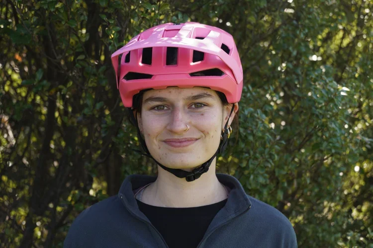 Person wearing a pink bicycle helmet smiling outdoors with green foliage background.