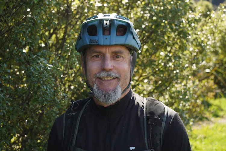 Smiling man wearing a blue bike helmet and black outdoor jacket, standing in a wooded area with sunlight filtering through the trees.