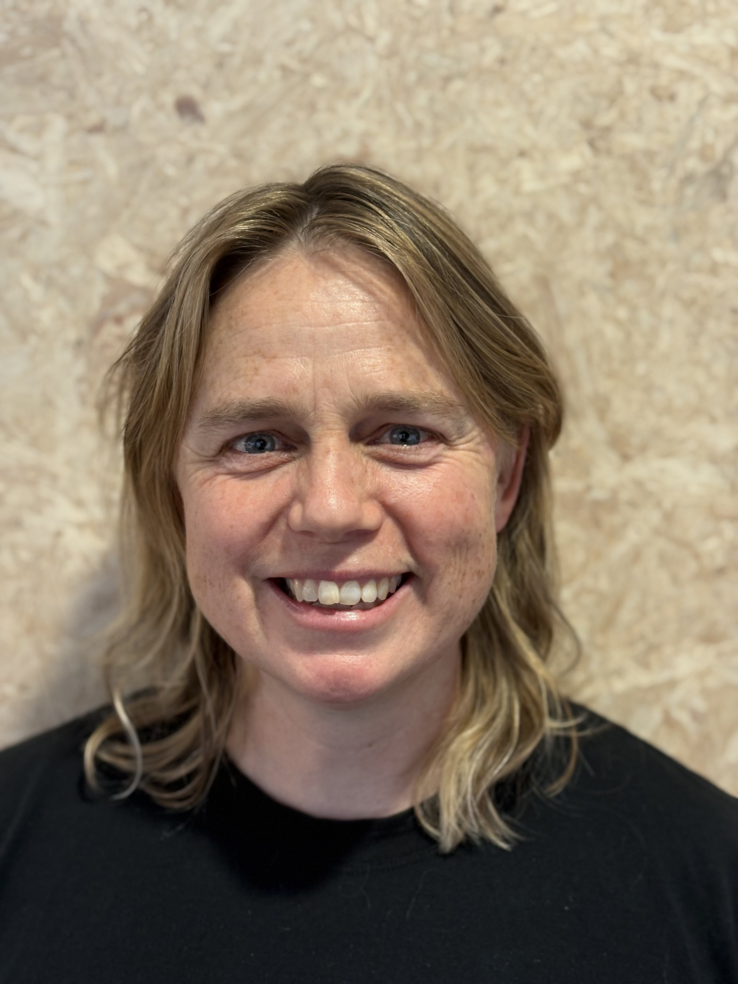 Close-up of a smiling woman with blonde hair, blue eyes, and freckles, wearing a black top, standing against a beige background.