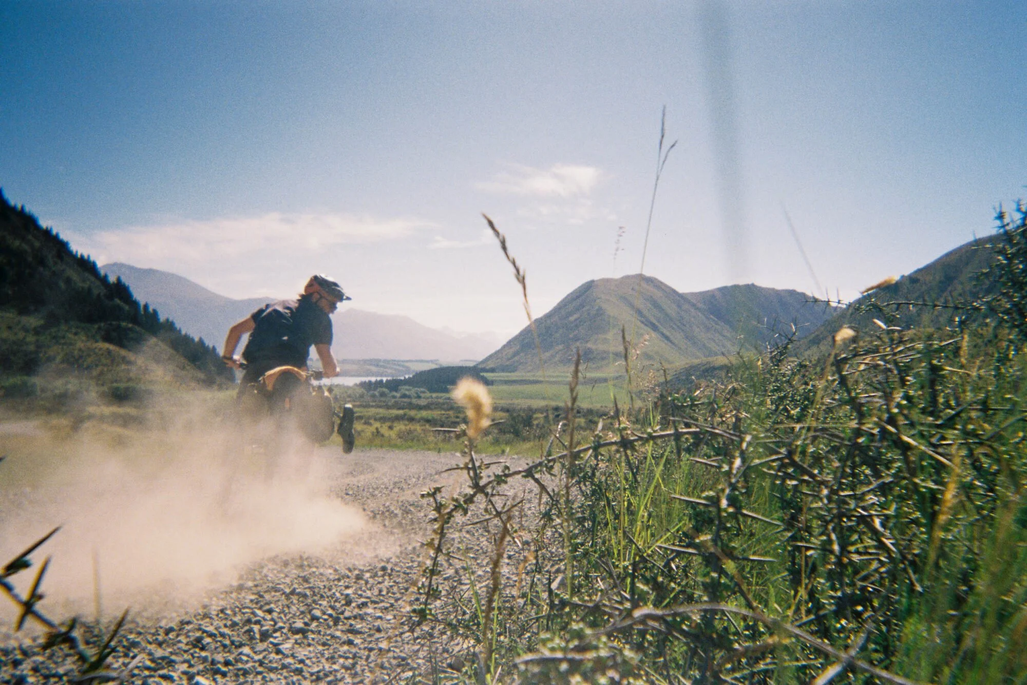 A person riding a dirt bike on a dusty trail in a mountainous landscape with a river, green hills, and a partly cloudy sky.
