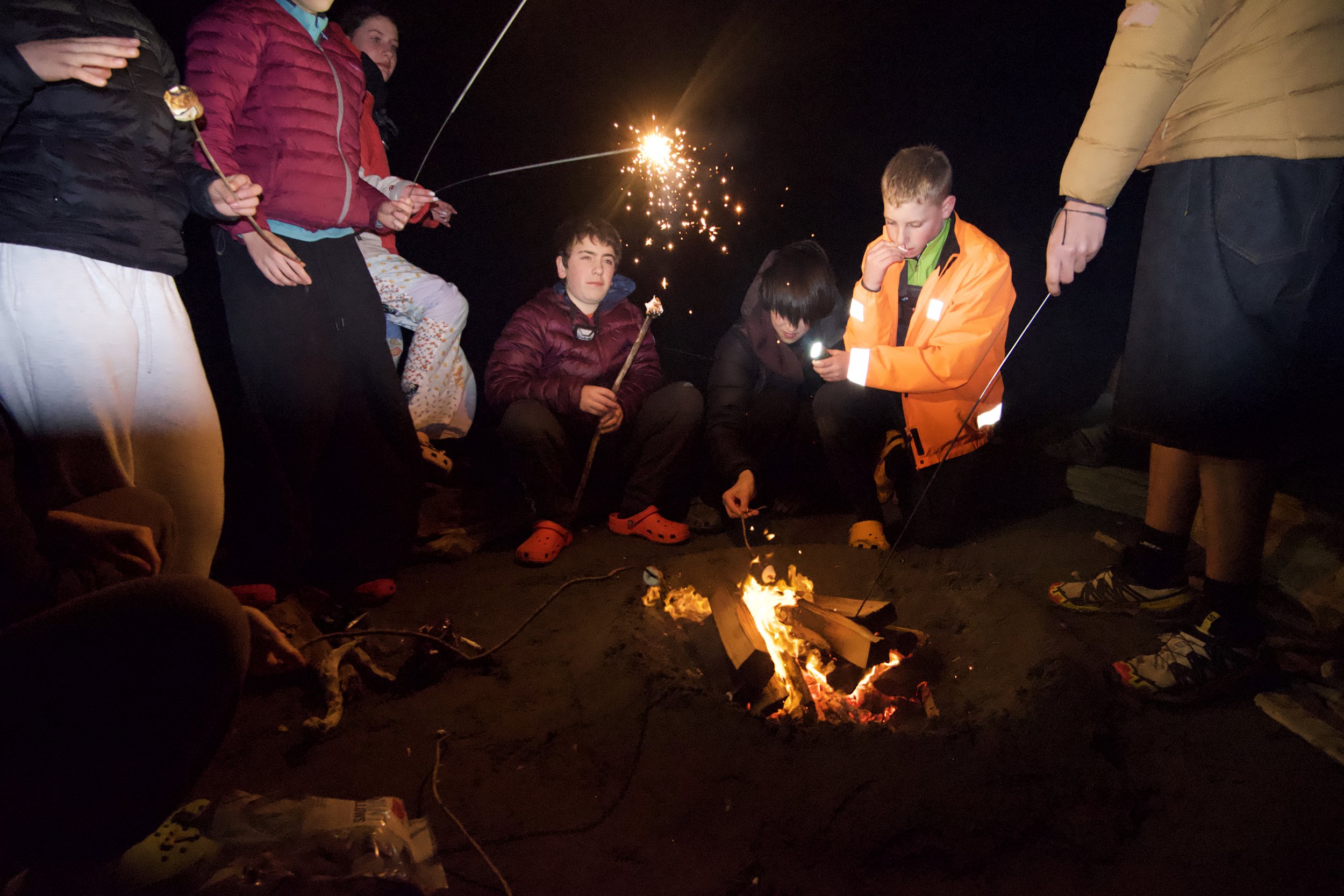 Group of people sitting and standing around a campfire at night, some holding sparklers, dressed in warm clothing.