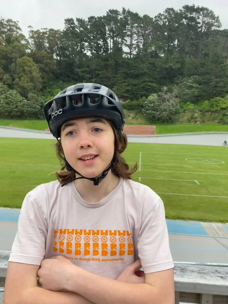 A young person wearing a safety helmet with arms crossed, standing at an outdoor sports stadium with a grass track and field area, surrounded by trees.