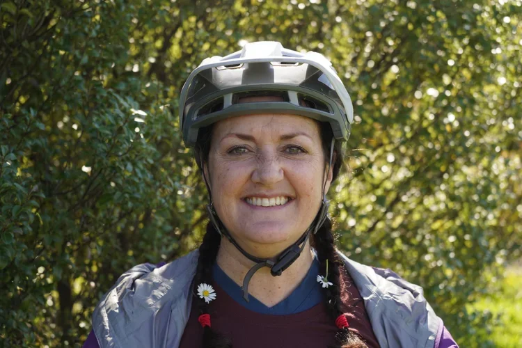 A smiling woman wearing a bicycle helmet, outdoors with green trees in the background.