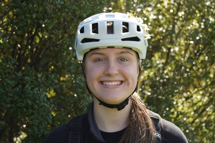 Young woman smiling, wearing a white bicycle helmet and black outdoor clothing, standing outdoors with green foliage in the background.