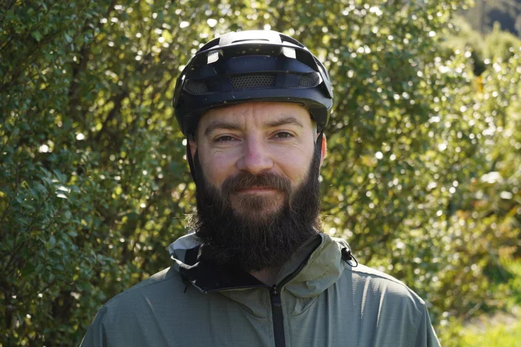 A man with a beard wearing a black helmet and a green jacket, outdoors with greenery in the background.