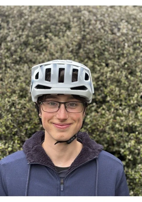 Young person wearing glasses, a white bicycle helmet, and a dark blue zip-up jacket with a black hoodie underneath, standing outdoors in front of a bushy background.