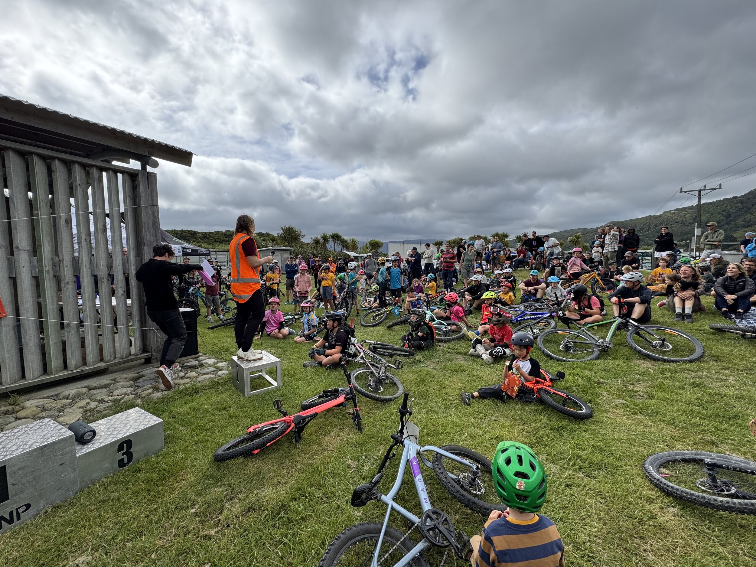 A large group of children and adults gathered outdoors on a cloudy day, many children wearing helmets with some sitting on their bicycles and others standing or sitting on the grass. There is a structure on the left side with a person in a safety vest speaking to the crowd, and a mountain landscape and power lines in the background.