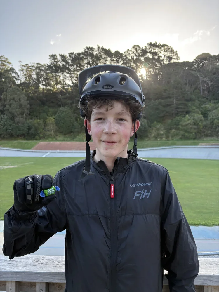A boy wearing a black helmet and black jacket, holding a small green object, standing outdoors on a wooden platform with a grassy field and trees in the background during sunset.