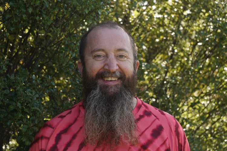 A man with a long beard and a red tie-dye shirt smiling outdoors with green foliage in the background.